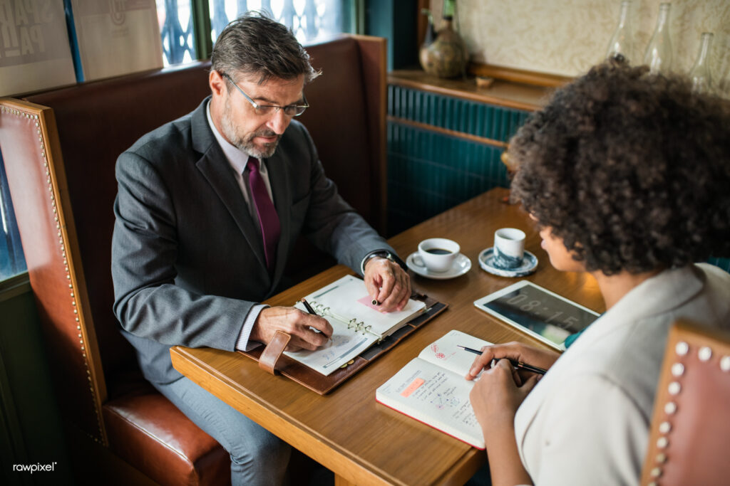 Two businesspeople sit across a café table with notebooks, a tablet, and coffee cups.